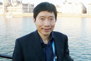 This photo is of Dr. Tim Liao. He is wearing a black sports coat over a navy blue shirt and standing in front of a river with buildings visible on the opposite bank.