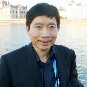 This photo is of Dr. Tim Liao. He is wearing a black sports coat over a navy blue shirt and standing in front of a river with buildings visible on the opposite bank.